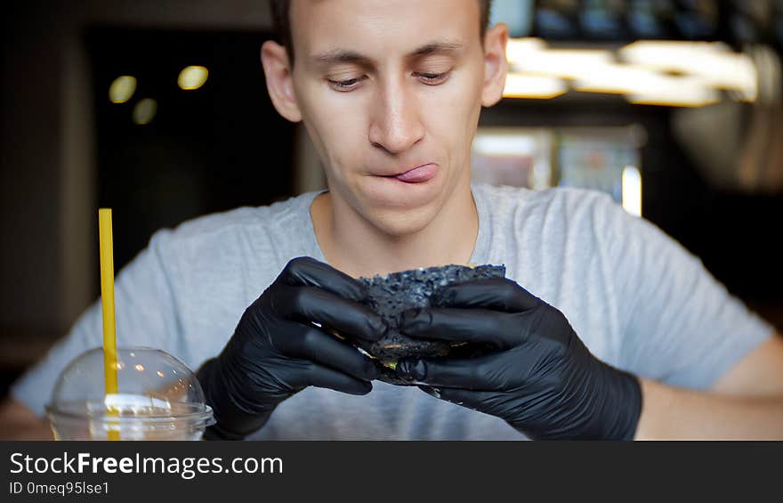 A young man sits in a restaurant and looks carefully at the burger who holds in his hands.