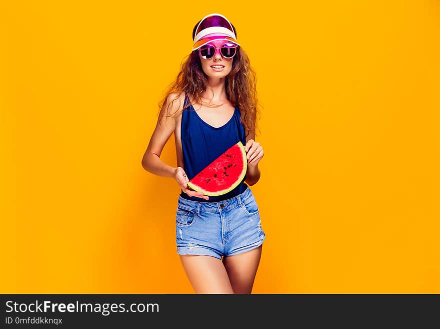 Beautiful young female in cap, sunglasses and swimsuit hold slice of watermelon and looking away while standing on yellow background. Copyspase. Beautiful young female in cap, sunglasses and swimsuit hold slice of watermelon and looking away while standing on yellow background. Copyspase