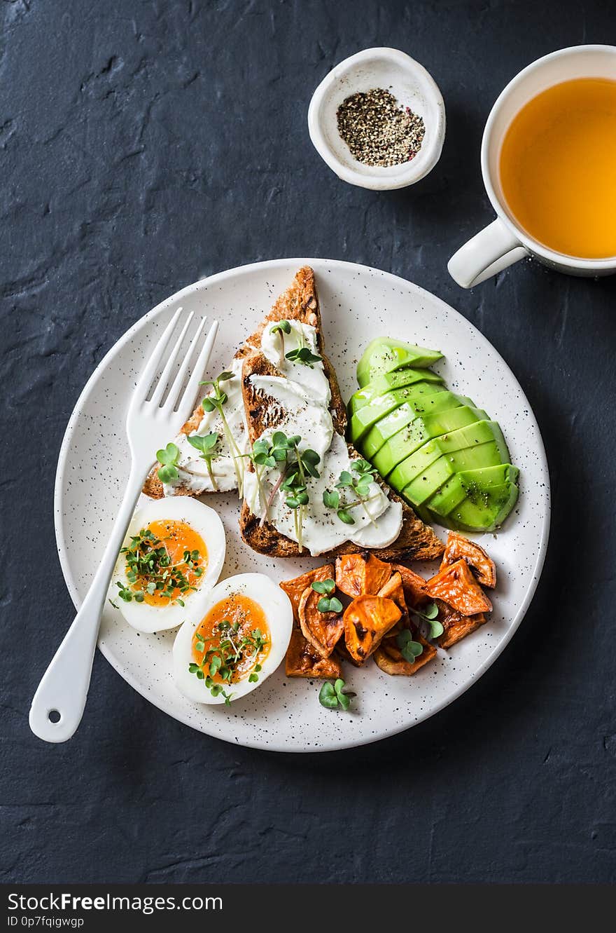 Cream cheese toast, avocado, boiled egg, baked sweet potatoes and tea - delicious healthy breakfast snack on a dark background