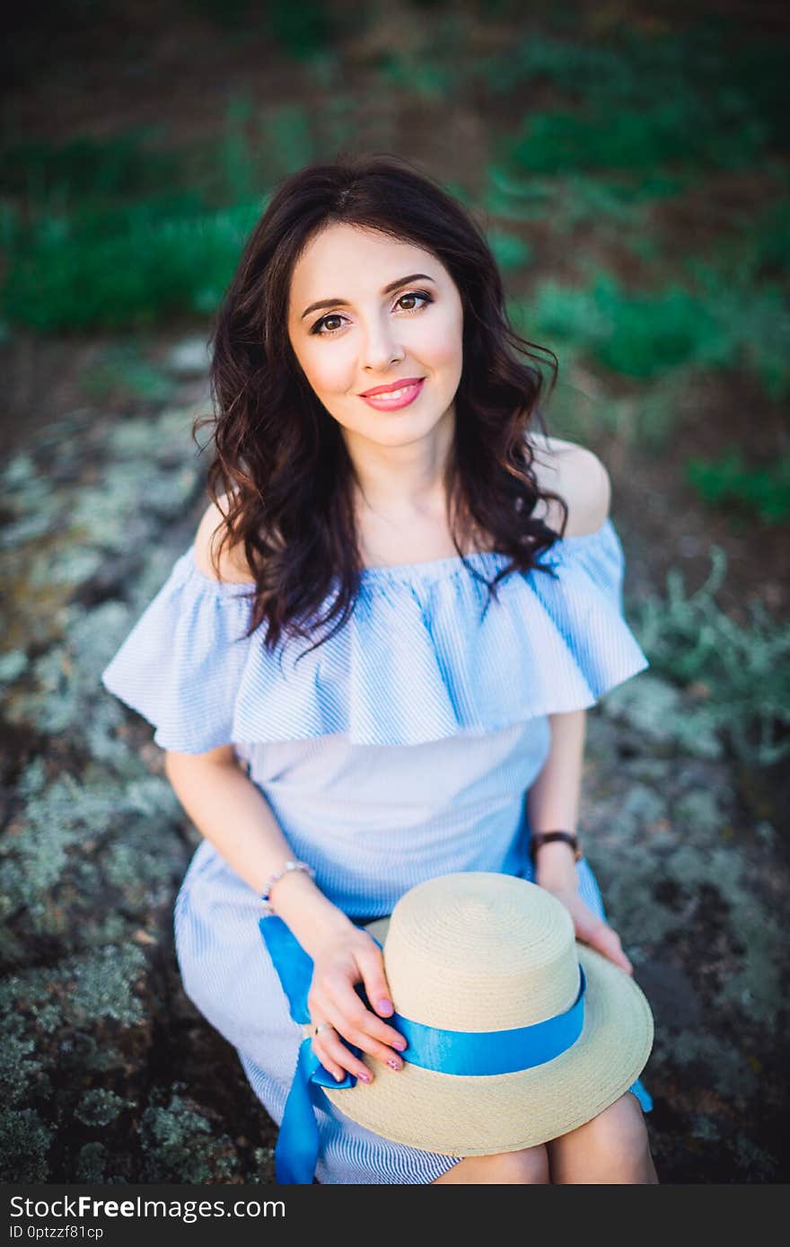 Close up Portrait of young woman in blue romantic dress with hat sitting on the stones. Calm and harmony. Summer vacation,fun, positive mood. Back sun light, sun beams. Vertical card. Copy space