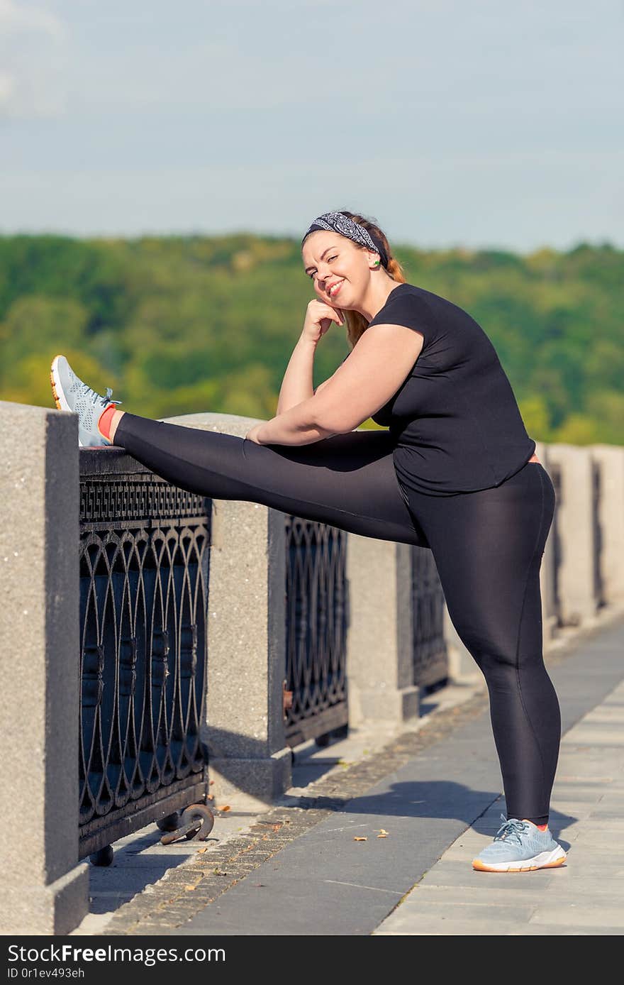 Young woman oversized doing stretching exercises on the embankment in a city park in the early morning