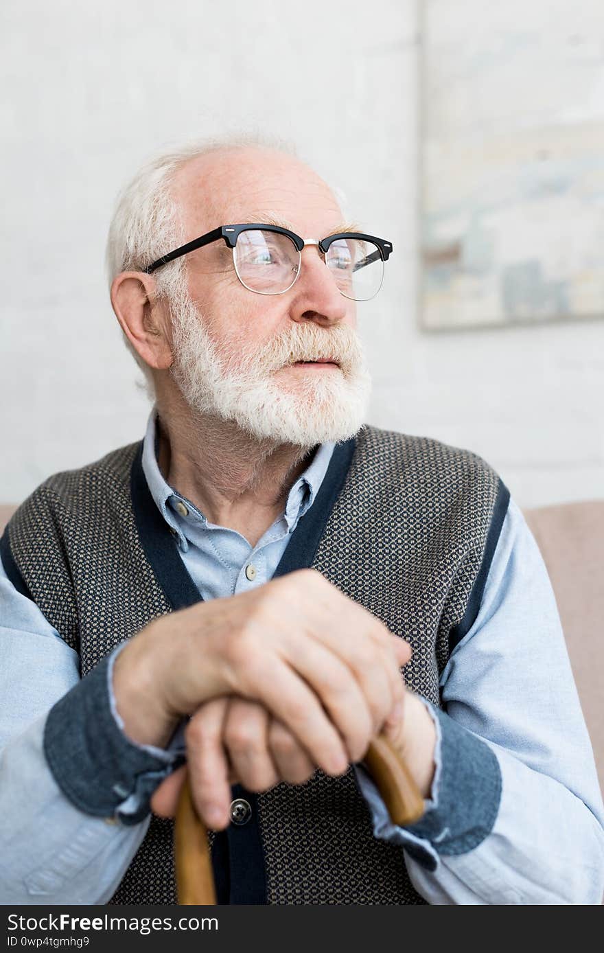 Calm and sad senior man with walking stick looking away, sitting in bright room