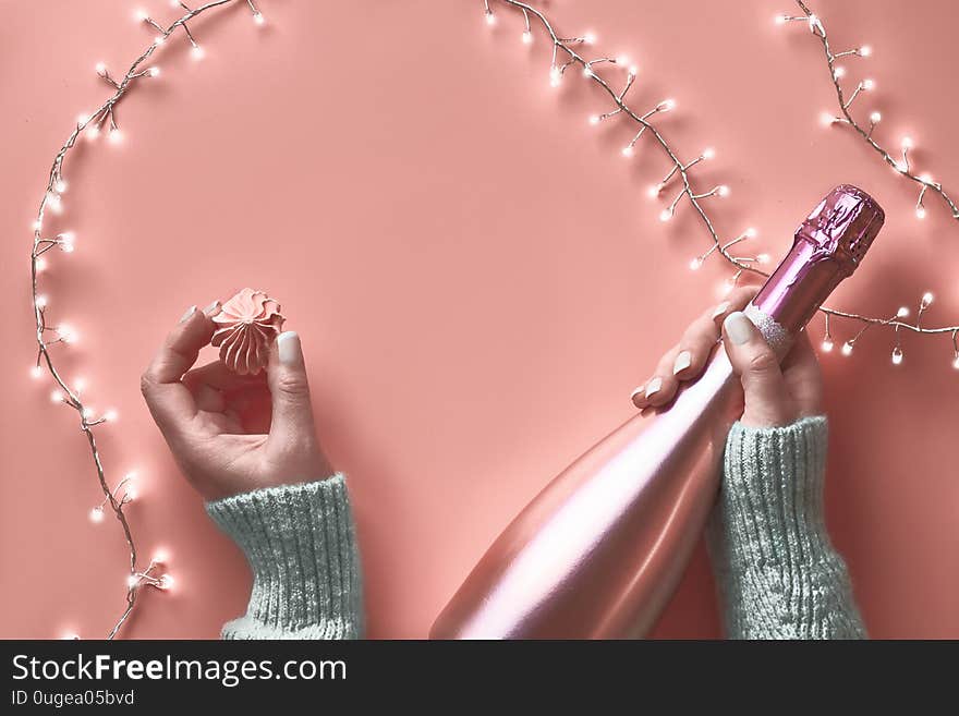 Valentine top view on pink background. Light garland, woman hands with metallic pink champagne bottle and sweet marshmallow snack