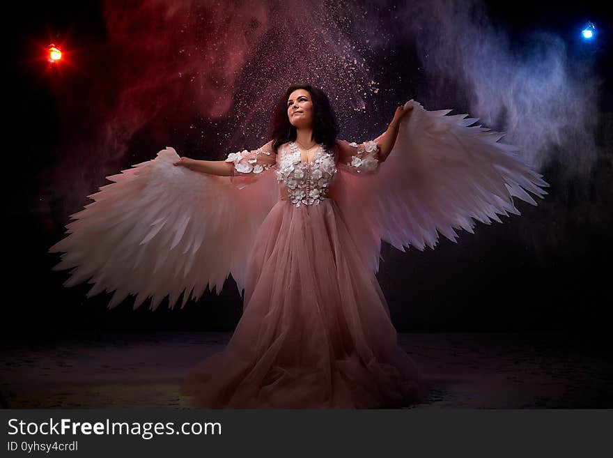 Girl with white wings posing in dark black studo during photoshoot with flour or dust and light. White angel in dark space