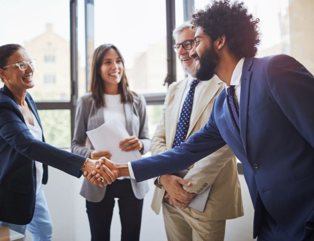 Two individuals shaking hands while two others are smiling and holding documents