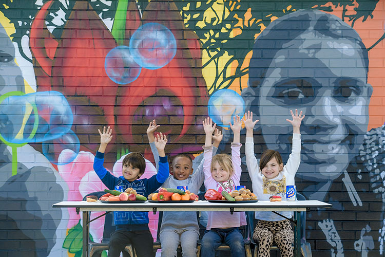 Four children with their arms up smile with fruit in front of them.