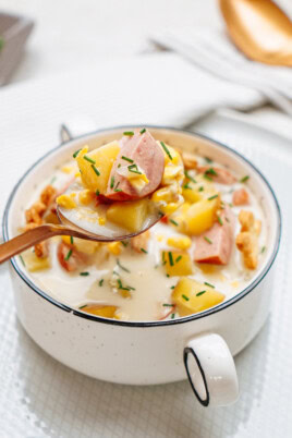 A bowl of creamy sausage corn chowder containing chunks of sausage, potatoes, and garnished with chopped chives. A spoon holding some of the soup is above the bowl.