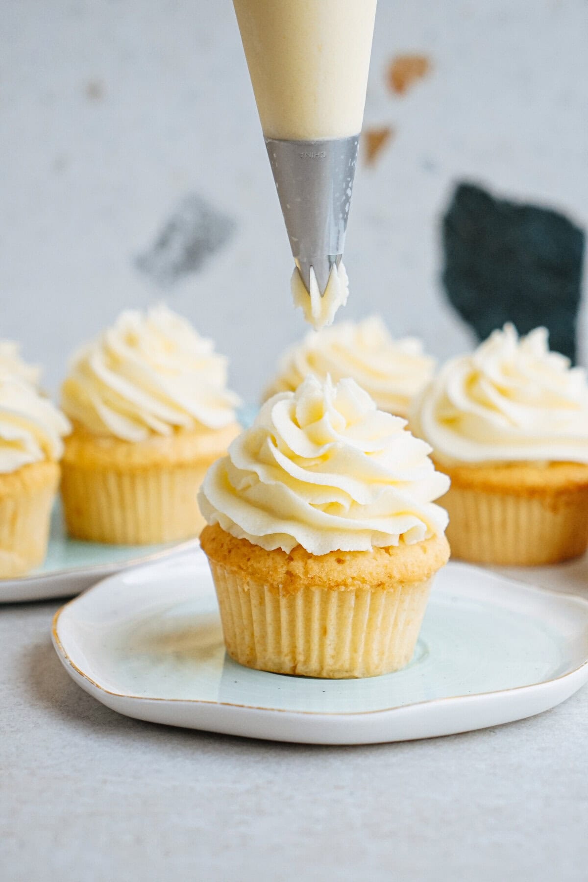 Piping frosting onto vanilla cupcakes arranged on a light blue plate.