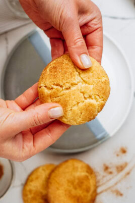 Hands breaking a round, snickerdoodle cookie over a plate, with more cookies and cinnamon nearby.