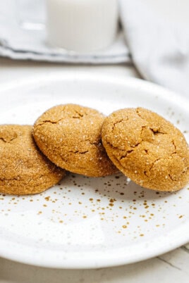 Three sugar-coated ginger cookies on a white speckled plate, with a blurred background featuring a glass of milk.