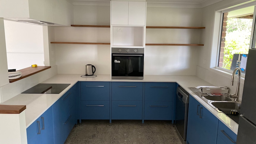 A sparse new kitchen, with blue cupboards , white bench top, polished concrete floor and wooden shelving.