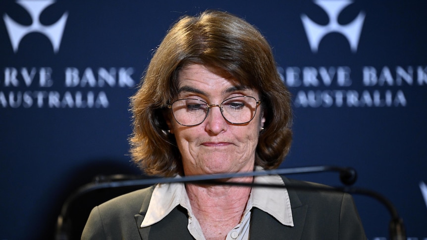 A woman with short brown hair and glasses looking down at her notes behind a lectern with two microphones.