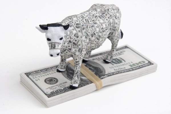 A cow covered in hundred dollar bills and standing on a stack of more hundred dollar bills.