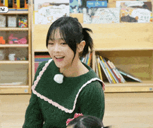 a woman in a green sweater smiles in front of a bookshelf with a book titled ' a rabbit ' on it