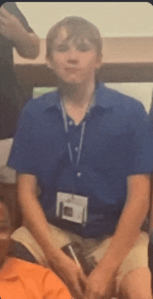 a young man wearing a blue shirt and lanyard is sitting in a room with other people .