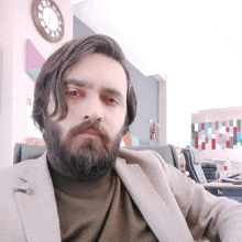 a man with a beard looks at the camera with a clock on the wall behind him