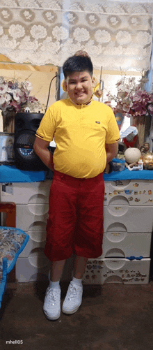 a young boy wearing a yellow shirt and red shorts stands in front of a dresser