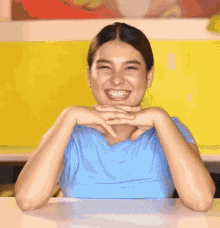 a woman in a blue shirt is sitting at a table with her hands on her chin and smiling .