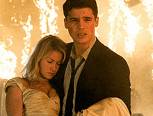 a man in a suit and tie is carrying a woman in a white dress in front of a fire .