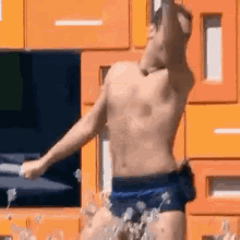 a shirtless man in blue swim trunks is dancing in front of a wall of orange drawers .