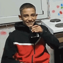 a young boy in a red and black jacket is sitting in front of a stove drinking from a glass .