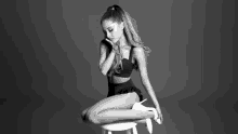 a black and white photo of ariana grande sitting on a stool with her legs crossed .