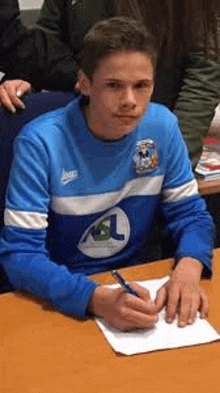 a young man in a blue shirt is sitting at a table writing on a piece of paper with a pen .