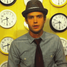 a man wearing a hat and tie stands in front of a wall of clocks including one that shows the time as 10:15