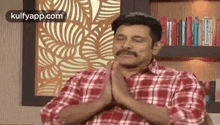 a man in a plaid shirt is sitting in front of a bookshelf with his hands folded in prayer .