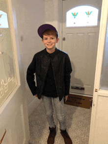 a boy wearing a purple hat and a black jacket stands in front of a door with a welcome mat