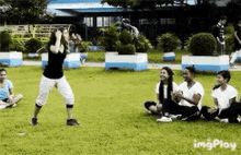 a group of people are sitting on the grass in a park while a woman stands in the middle of the field .