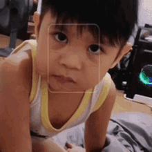 a young boy wearing a yellow and white tank top is sitting on a bed looking at the camera .