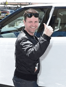 a man wearing sunglasses is giving a thumbs up while standing next to a white car .