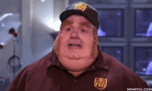 a fat man wearing a fbd hat and a brown shirt is standing in front of a wall of televisions .