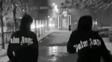 a black and white photo of two people wearing palm angels hoodies walking down a snowy street .