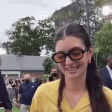 a woman wearing glasses and a yellow shirt is smiling while standing in front of a crowd of people .