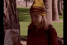 a young girl wearing a yellow and red striped hat is standing next to a tree holding a piece of paper .