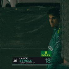 a man wearing a green rolex shirt stands in front of a black curtain