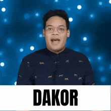 a man wearing glasses and a blue shirt stands in front of a banner that says dakor