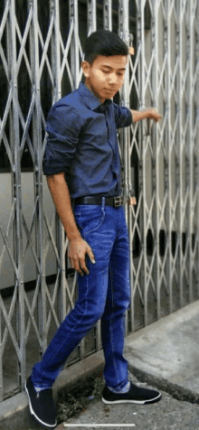 a young man in a blue shirt and blue jeans is standing in front of a fence