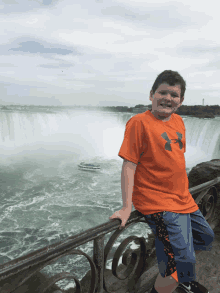 a boy wearing an orange under armour shirt stands on a railing overlooking a waterfall