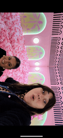 two women standing next to each other in front of a wall filled with pink balls