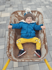 a young boy in a blue jacket and yellow pants is sitting in a wheelbarrow