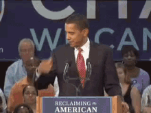 a man in a suit and tie stands at a podium with a sign that says " reclaiming the american "