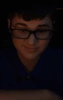 a young man wearing glasses and a blue shirt smiles in the dark