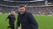 a man in a black jacket is running on a soccer field in front of a sign that says spell