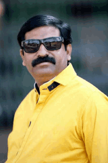 a man with a mustache wearing sunglasses and a yellow shirt is looking at the camera .