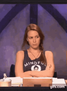 a woman is sitting at a table with her arms crossed and wearing a tank top that says mindful .