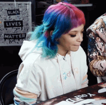 a woman with blue and pink hair sits in front of a sign that says " black lives matter "