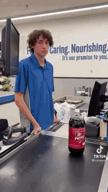 a man in a blue shirt is standing at a cash register next to a bottle of dr. perkis soda .